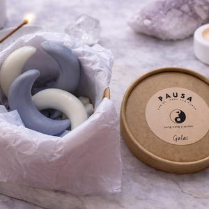 Puede incluir: Una caja con jabones en forma de media luna en tonos blancos y azules, envueltos en papel de seda. Junto a ella, un recipiente redondo con la etiqueta "PAUSA" y "Galai". También se ven una vela encendida y un incienso.