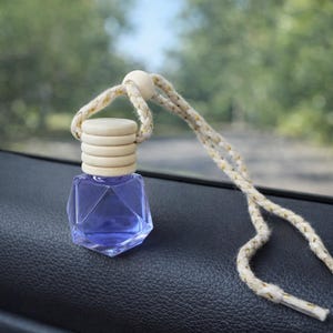 Puede incluir: Una pequeña botella de vidrio facetado llena de líquido azul, coronada con una tapa de madera y una cuerda color crema. La botella cuelga dentro de un coche, con un fondo verde borroso.