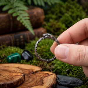 May include: A silver-colored ring in the shape of a snake biting its own tail. The ring has a textured surface, and the snake's head is detailed. The ring is held against a backdrop of moss, wood, and books.