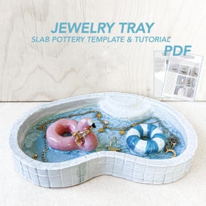 May include: A ceramic jewellery tray shaped like a swimming pool, with a pink flamingo and a blue and white inflatable ring. The tray has a light blue interior and is decorated with gold jewellery. The text "JEWELRY TRAY" is visible.