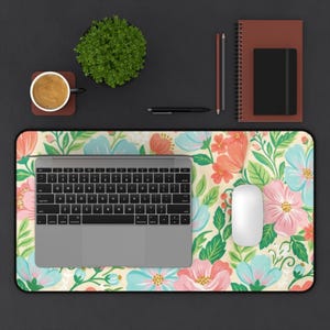 Puede incluir: Un mousepad floral con una computadora portátil, un ratón y otros accesorios de escritorio. El mousepad presenta un fondo crema con flores rosas, azules y naranjas. También hay una taza de café, una planta y un cuaderno.