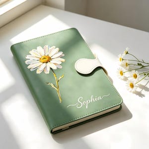 Pode incluir: Um caderno verde-sálvia com uma ilustração de uma margarida branca na capa. O caderno inclui uma aba de fecho magnético branca e o nome Sophia impresso em letra cursiva branca no canto inferior direito da capa.