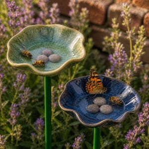 Pode incluir: Duas bacias de água de cerâmica em forma de flor em estacas de metal verde. A bacia superior é verde-clara com esmalte craquelado, contendo água, três pedras cinzentas lisas e duas abelhas. A bacia inferior é azul-escura com uma borboleta.