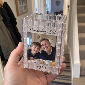 Op de afbeelding: Een doorzichtige, rechthoekige fotolijst gemaakt van bouwstenen met een foto van een kind en een volwassene. Het bovenste gedeelte bevat de tekst "Keep Smiling, Dad" en het onderste gedeelte de naam "Sophie" in een zwart cursief lettertype.
