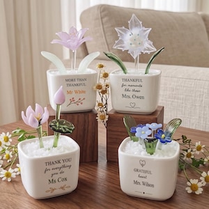 May include: Four white square ceramic planters filled with white pebbles, each containing a unique glass flower sculpture. The planters feature printed text including names like Mr. White, Mrs. Owen, Ms. Cox, and Mrs. Wilson, alongside messages of gratitude.