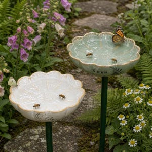 Könnte beinhalten: Zwei Keramik-Gartenstecker mit gewellten, blütenförmigen Schalen. Eine Schale ist hellgrün mit dem Namen Sienna und einem Farn-Motiv, die andere weiß mit dem Namen Lily und einem Mond-Motiv. Beide enthalten Wasser, Bienen und Schmetterlinge.