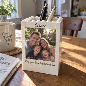 Peut inclure: Un porte-stylo rectangulaire blanc construit à partir de blocs de construction en plastique emboîtables. L'avant présente une photographie imprimée d'une famille avec le texte "Grace" en haut et "May your home be filled with love" en bas.