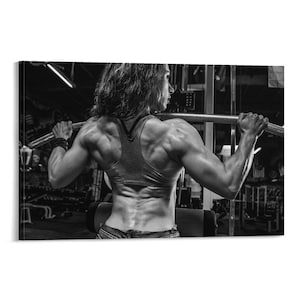Op de afbeelding: Zwart-wit foto van een persoon die in een sportschool traint. De persoon, gekleed in een grijze tanktop en spijkerbroek, trekt aan een stang van een gewichthefmachine. De rugspieren zijn zichtbaar.