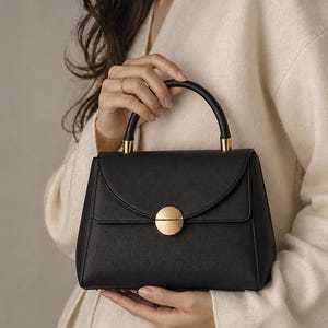 May include: Black handbag with a curved top flap and a gold-coloured circular clasp. The bag has a rounded handle and gold-coloured accents. The bag is held by a person wearing a cream-coloured sweater.