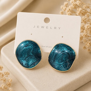 May include: A pair of teal and gold earrings. The earrings have a marbled teal design with a gold border. The earrings are displayed on a white jewelry card with the word "JEWELRY" printed on it. The card also says "Silver 925 post".