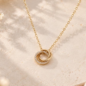 May include: A gold-toned necklace featuring a pendant composed of two interlocking circular rings. One ring is plain polished gold, while the other is encrusted with small, clear, sparkling stones. The pendant hangs from a delicate gold-toned chain.
