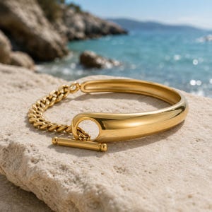 May include: A gold-toned bracelet featuring a polished, smooth semi-circular band connected to a textured curb chain. The piece is secured with a toggle clasp mechanism, consisting of a horizontal bar that slides through a circular loop.
