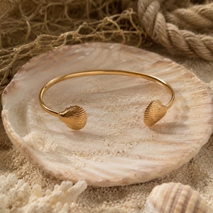 Op de afbeelding: Een goudkleurige open armband met twee kleine, getextureerde schelpvormige uiteinden. De metalen band is glad en cirkelvormig, ontworpen om om de pols te dragen waarbij de twee schelpaccenten symmetrisch naar voren zijn gericht.