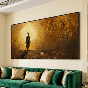 Puede incluir: Obra de arte enmarcada de gran tamaño que representa una silueta frente a una luz brillante, con antiguos jeroglíficos en el fondo. La pintura es en tonos dorados y marrones, con un marco negro. Cuelga sobre un sofá de terciopelo verde con cojines decorativos.