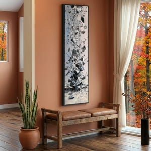 May include: A framed vertical abstract artwork in a black frame. The art shows a grayscale explosion of fragmented shapes against a light background. A wooden bench with leather cushions is below the artwork. A potted plant and a window with autumn foliage are also visible.