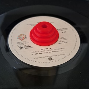 May include: A red record stabilizer sits on a black vinyl record. The record label displays the song title "Whip It" and the album title "Freedom of Choice". The stabilizer is a tiered, cylindrical shape.
