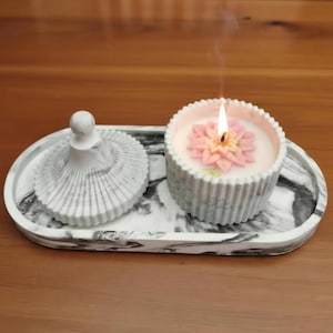 Pode incluir: Uma vela acesa com um desenho de flor rosa está em uma bandeja com padrão de mármore, ao lado de um recipiente com tampa combinando. A vela está em um recipiente cinza canelado. A bandeja e o recipiente têm um design de mármore branco e cinza.