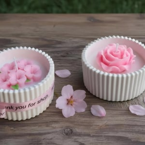 Peut inclure: Deux bougeoirs blancs et côtelés avec des bougies florales roses. Une bougie en forme de rose, l'autre avec plusieurs boutons floraux. Un ruban rose avec du texte entoure un bougeoir. Des pétales de fleurs et une pierre grise sont sur la surface en bois.