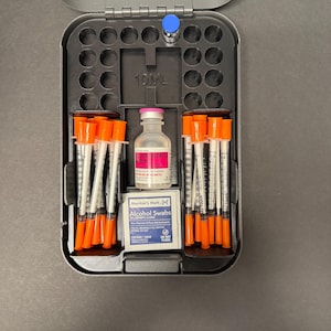 May include: An open, gray medical case containing syringes with orange caps, a vial with a pink label, and a small box of alcohol swabs. A small blue-capped vial is also visible. The case is on a dark surface.