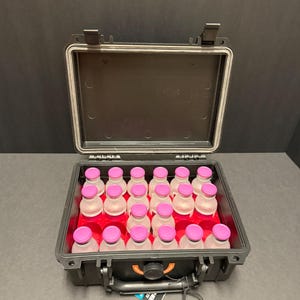 May include: An open black hard case with a red interior holds multiple clear bottles with pink caps. The case is on a dark surface. The bottles are arranged in rows.