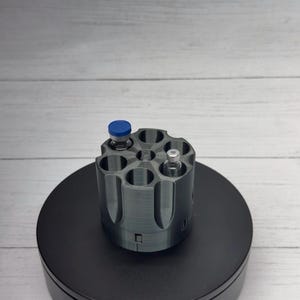 May include: A gray, cylindrical, six-chambered revolver cylinder holder designed to store small vials. One chamber contains a vial with a blue cap, and another contains a vial with a silver cap. The object is positioned on a circular black base.