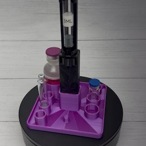 May include: A purple 3D-printed square organizer stand with multiple circular slots for vials. A black vertical holder is centered on the base, containing a clear glass syringe with a 3ML label. Two small glass vials are placed in the slots.
