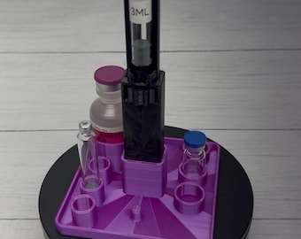 May include: A purple 3D-printed square organizer stand with multiple circular slots for vials. A black vertical holder is centered on the base, containing a clear glass syringe with a 3ML label. Two small glass vials are placed in the slots.