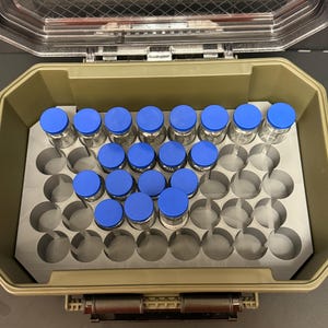 Könnte beinhalten: Ein olivgrüner Hartschalen-Aufbewahrungskoffer mit transparentem Deckel und einer grauen Schaumstoffeinlage mit kreisförmigen Aussparungen. Mehrere kleine, durchsichtige Glasfläschchen mit leuchtend blauen Kunststoffkappen stecken in den Schaumstoffschlitzen.