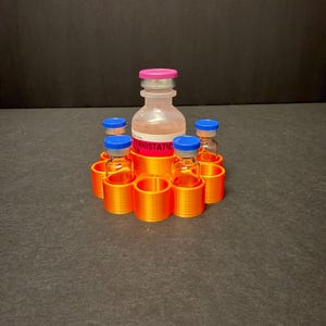 May include: An orange 3D-printed holder with six circular slots, each designed to hold a small vial. A larger vial with a pink cap sits in the center. The vials have blue caps. The central vial has the word "ERIOSTATIC" printed on it.