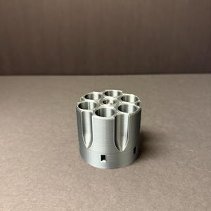 Puede incluir: Un objeto cilíndrico de color plateado con múltiples aberturas circulares. El objeto tiene una serie de ranuras verticales a lo largo de su exterior y parece estar hecho de metal. El diseño sugiere un componente mecánico o industrial.