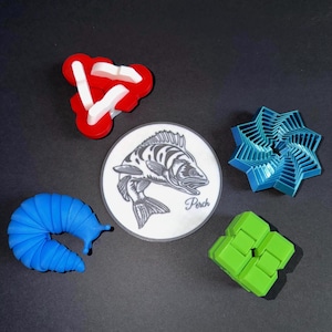 May include: An assortment of colorful 3D-printed objects on a black surface. Includes a red and white geometric shape, a blue slug, a blue starburst, a green cube, and a circular graphic of a fish with the word "Perch".