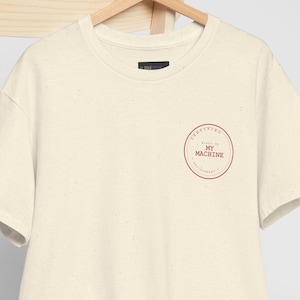 May include: A cream-coloured short-sleeve t-shirt with small dark speckles and a circular graphic on the chest containing the text "MADE IN MY MACHINE". A plain white baseball cap is draped over the back of the wooden chair holding the shirt.