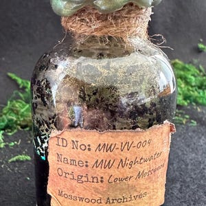 May include: A small, clear glass bottle with a green, iridescent stopper. The bottle is filled with a dark liquid and has a label that reads "MW Nightwater" from "Mosswood Archives". The bottle is sealed with twine.