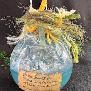 May include: A spherical glass jar with a blue and silver interior, adorned with a cluster of textured, multi-coloured fibres. The jar features a label stating "Spring Crystal Drift" and "Mosswood Archives."