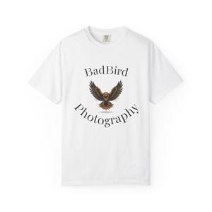 May include: White t-shirt featuring the text "BadBird Photography" in a curved font, with a detailed illustration of a bird with outstretched wings. The bird illustration has brown and gold tones.