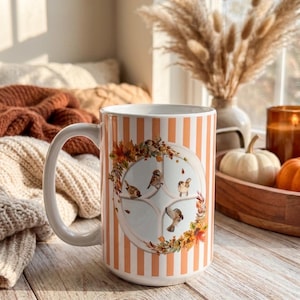 Puede incluir: Una taza de cerámica blanca con rayas verticales naranjas y un gráfico circular central. El gráfico muestra un marco de ventana rodeado por una corona de hojas de otoño y varios pájaros pequeños de color marrón posados o volando.