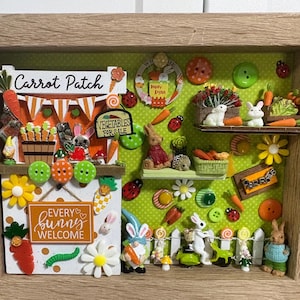 May include: A framed Easter-themed shadow box with a wooden frame. The box features miniature bunnies, carrots, flowers, and signs that read "Carrot Patch" and "Every Bunny Welcome". The background is green with decorative buttons.