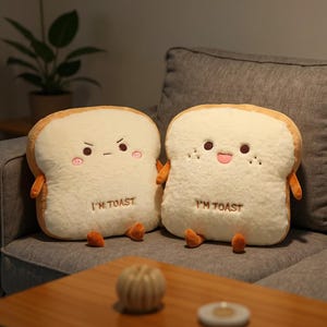 May include: Two plush bread slice pillows with cartoon faces and the text "I'M TOAST". The pillows have white centers, tan crusts, and small orange arms and feet. One has an angry expression, the other a happy one. They are on a gray couch.