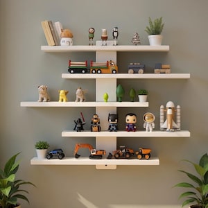 Op de afbeelding: Een witte wandplank met diverse decoratieve items. De planken bevatten kleine beeldjes, speelgoedvoertuigen, potplanten en boeken. De items omvatten een speelgoedvrachtwagen, een raket en superheldenfiguren.