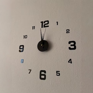 May include: A modern wall clock with black numerals and hands against a light-colored wall. The clock face is minimalist, with the numbers 1 through 12 individually placed. The clock hands point to 12 o'clock.