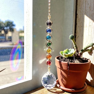 May include: A decorative suncatcher featuring a clear crystal ball and seven coloured beads in a gold-tone metal frame. The beads are arranged in a rainbow spectrum. A small potted succulent is also visible.