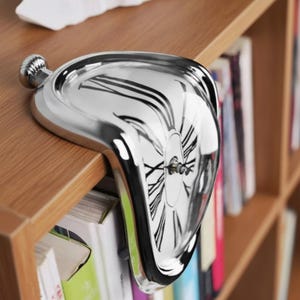 Puede incluir: Un reloj plateado y derretido con números romanos negros, apoyado sobre una estantería de madera. El reloj tiene una parte superior bulbosa y un diseño distorsionado y surrealista, que se asemeja a un reloj de bolsillo derritiéndose. Un artículo decorativo único.