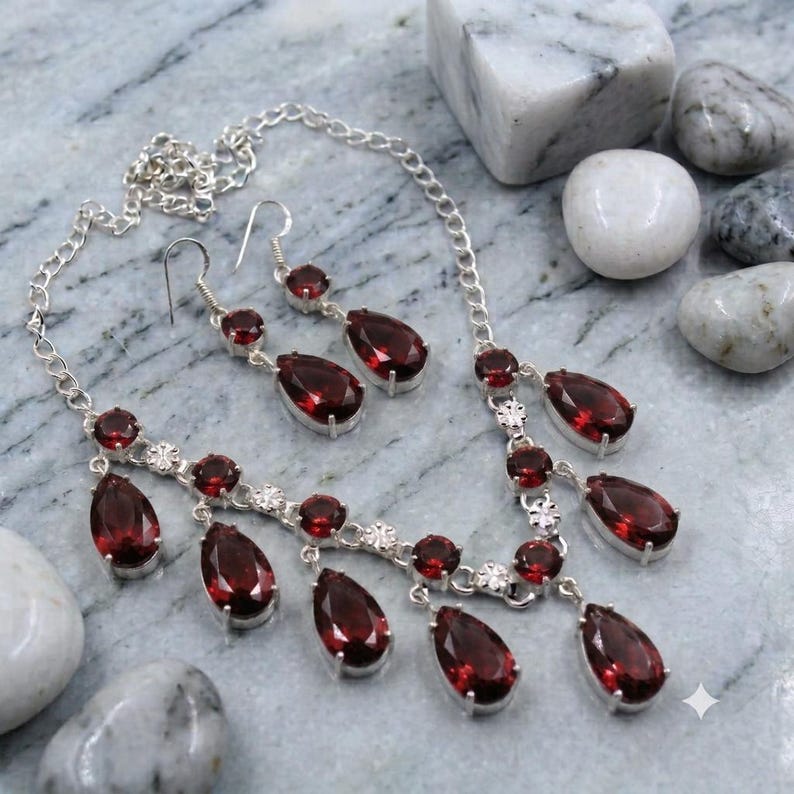 May include: A silver-toned necklace and matching pair of drop earrings featuring deep red, pear-shaped faceted stones. The necklace has a delicate chain with multiple dangling stone pendants, and the earrings have a single matching stone drop.