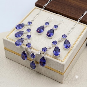 May include: A silver-toned jewelry set consisting of a necklace and matching drop earrings. The necklace features a chain with multiple dangling teardrop-shaped purple stones and small clear round stones. The earrings mirror this design.