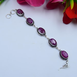 May include: A silver bracelet with oval, deep red gemstones. Each gem is set in a decorative bezel and linked by silver. The bracelet is shown against a white background with floral accents.