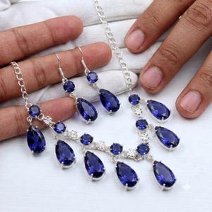 May include: A silver-toned jewelry set consisting of a necklace and a pair of drop earrings. The set features multiple deep blue, pear-shaped faceted stones and smaller round faceted stones set in silver-toned metal links with floral accents.
