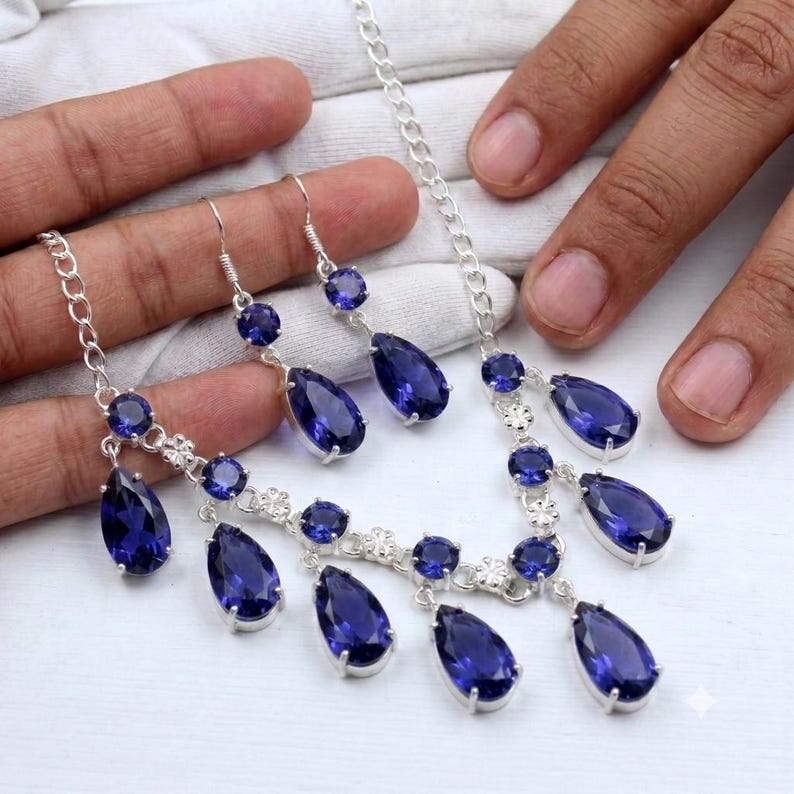 May include: A silver-toned jewelry set consisting of a necklace and a pair of drop earrings. The set features multiple deep blue, pear-shaped faceted stones and smaller round faceted stones set in silver-toned metal links with floral accents.
