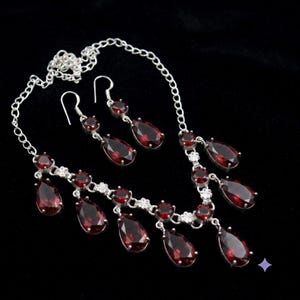 May include: A silver-toned necklace and matching pair of drop earrings featuring faceted, pear-shaped deep red garnet gemstones. The necklace chain is adorned with small round garnet stones and floral-shaped silver accents between the drops.