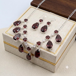 May include: A silver necklace and matching earring set featuring deep red, pear-shaped garnet gemstones. The necklace has a delicate silver chain with multiple dangling garnet drops, while the earrings consist of single pear-shaped garnet stones.
