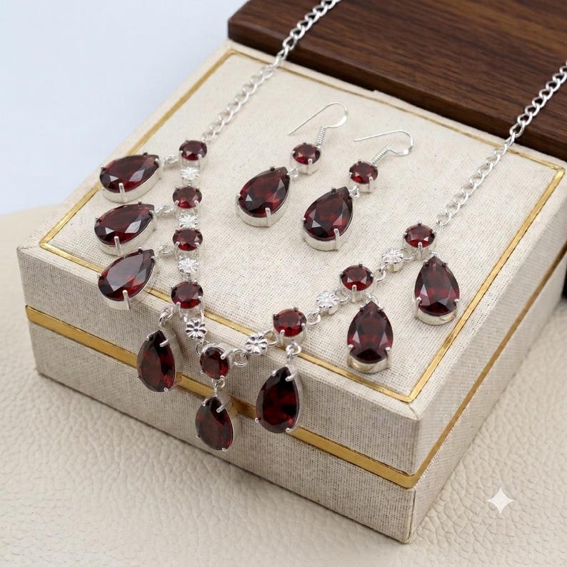 May include: A silver necklace and matching earring set featuring deep red, pear-shaped garnet gemstones. The necklace has a delicate silver chain with multiple dangling garnet drops, while the earrings consist of single pear-shaped garnet stones.